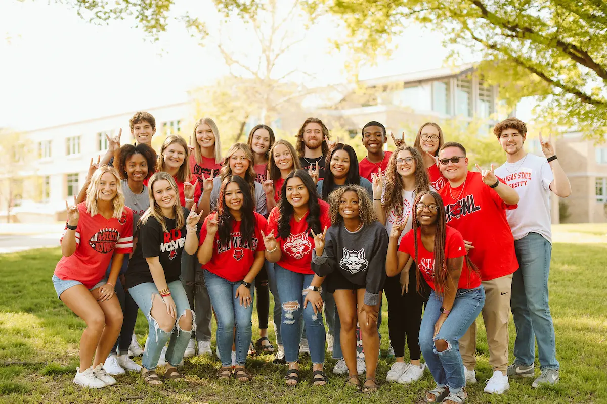 Diverse group of A-State students wearing Red Wolves gear gather outdoors, smiling and making the wolf hand sign.