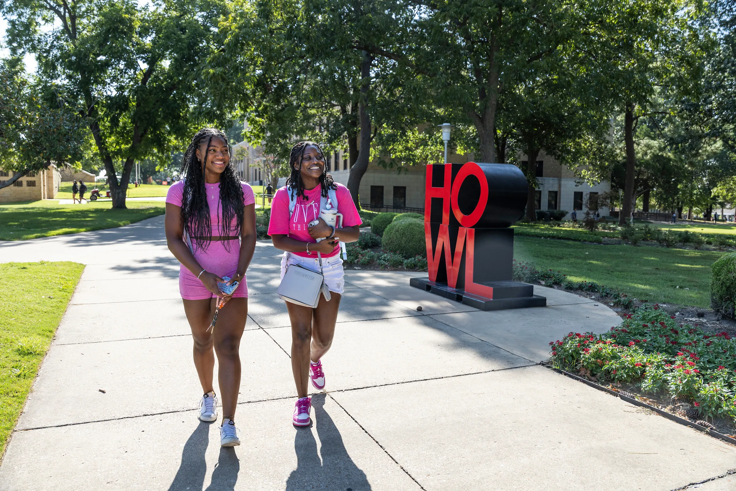 Two students in pink outfits walk past the HOWL sculpture on a sunny day at campus.