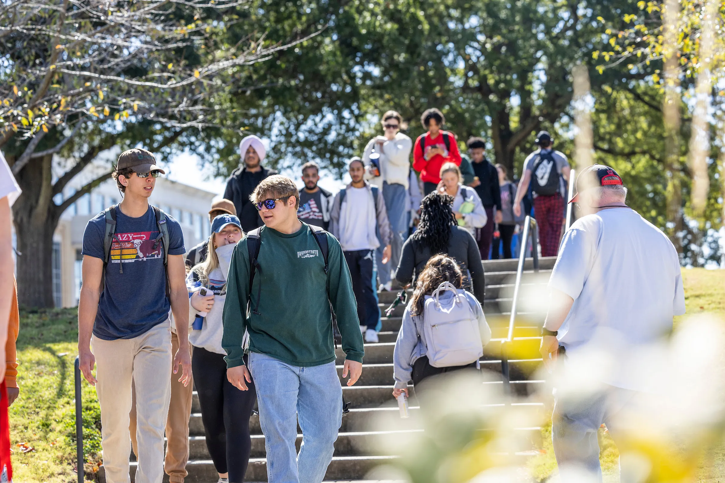 Large group of students walks down campus stairs on a bright day with fall leaves and backpacks.