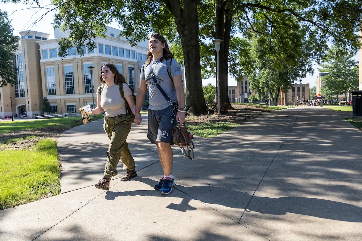 Two A-State students walk confidently down a sidewalk through the heart of campus.