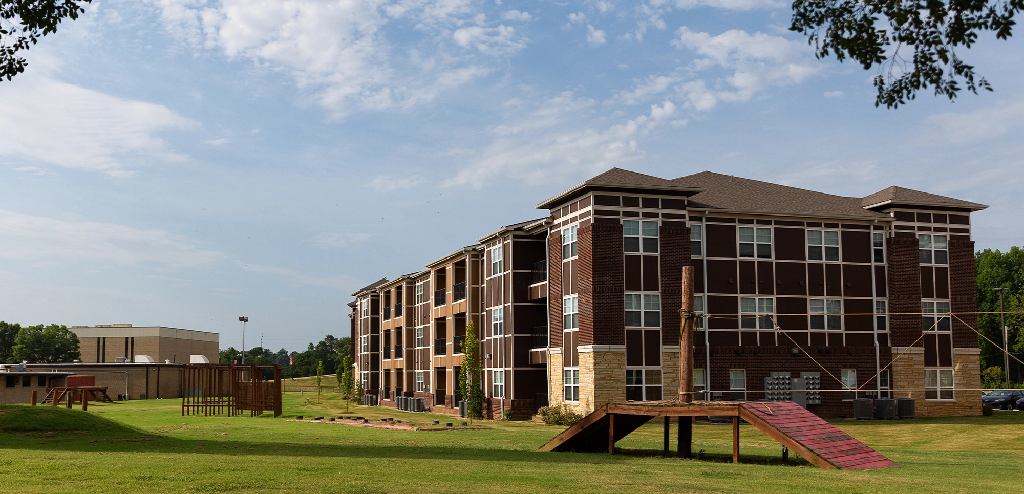 Exterior view of The Circle apartment complex showing a multi-story brick building with large windows, surrounded by green lawns and a wooden walkway under a partly cloudy sky.