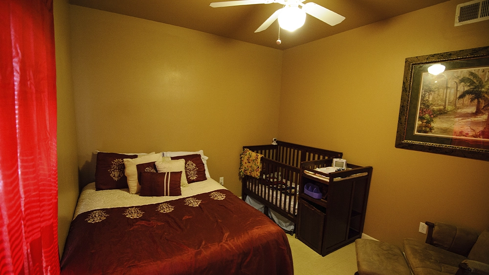 Bedroom with a full-size bed in burgundy bedding, a crib, and a dresser, accented by red curtains and framed wall art.
