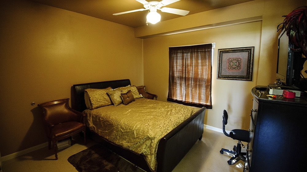 Bedroom with a full-size bed covered in gold bedding, two accent chairs, a dresser with TV, and a desk chair near a window with wooden blinds.