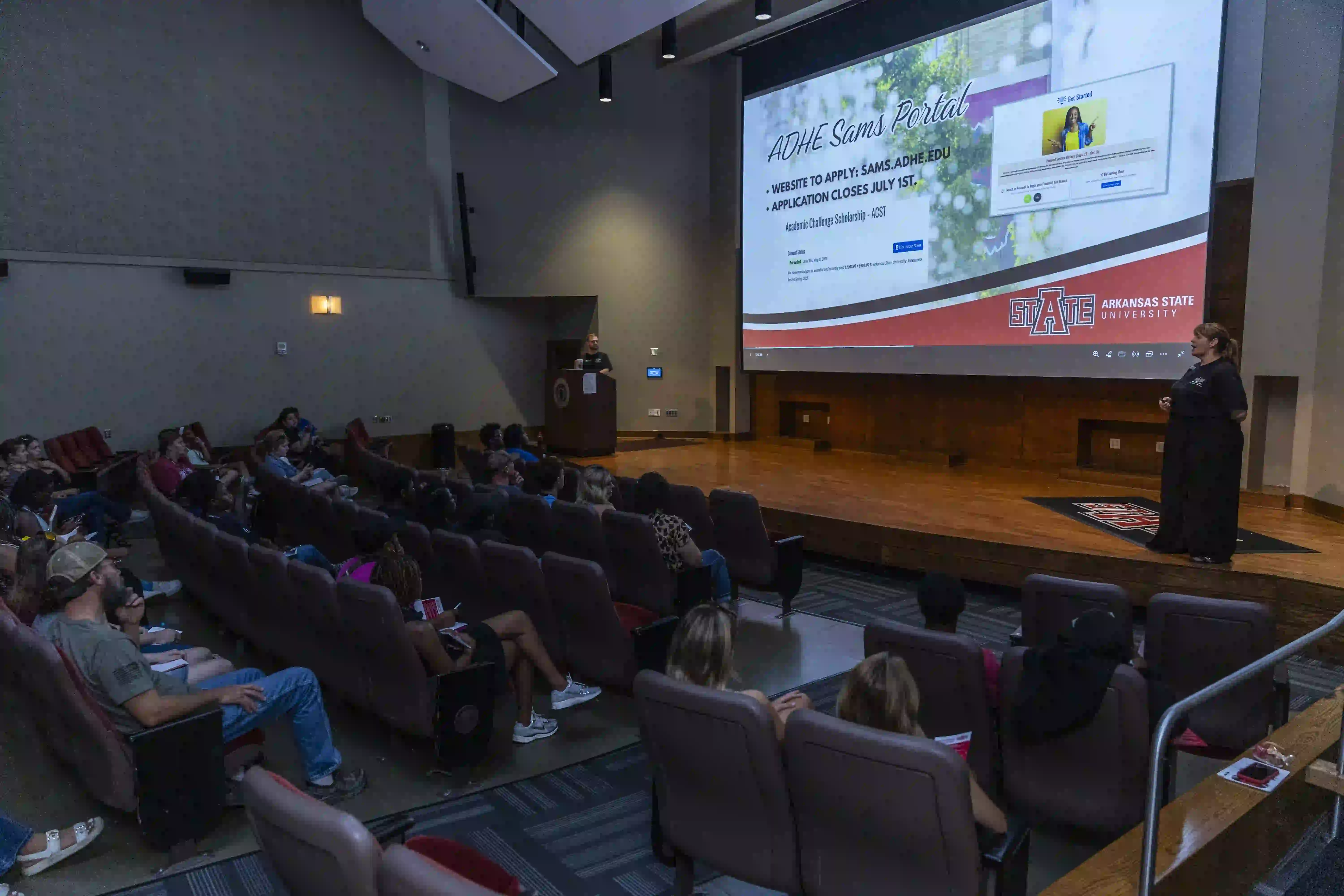 Speaker on stage giving presentation to students in auditorium.