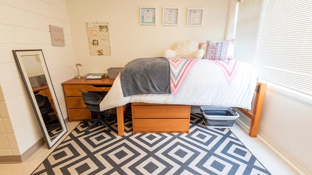 Bedroom view showing a lofted bed with colorful bedding, a wooden desk and chair, and a full-length mirror leaning against the wall.