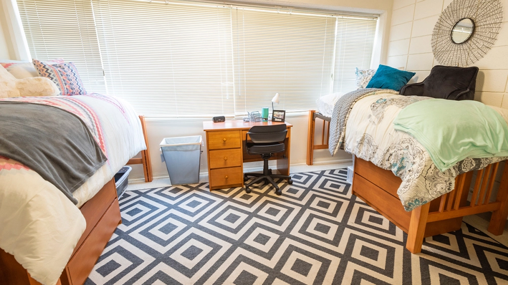 University Hall bedroom with two lofted beds, colorful bedding, and a shared desk positioned between the beds. Large windows with blinds allow natural light into the room, and a geometric rug covers the floor.