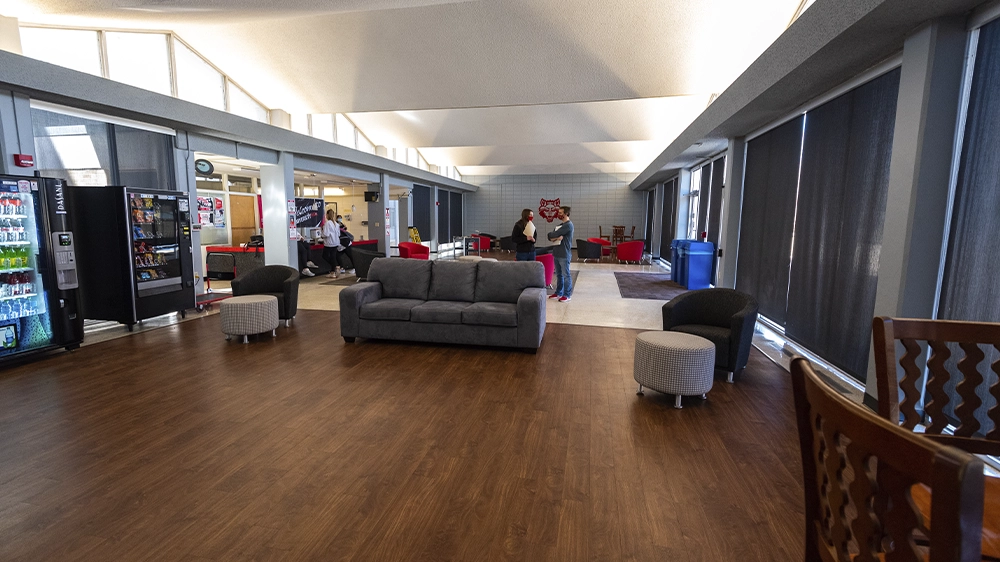 Spacious lobby inside University Hall featuring a gray sofa, round ottomans, vending machines, and large windows with black shades. Several people are visible near the back of the room, and the space includes bright overhead lighting and wood-style flooring.