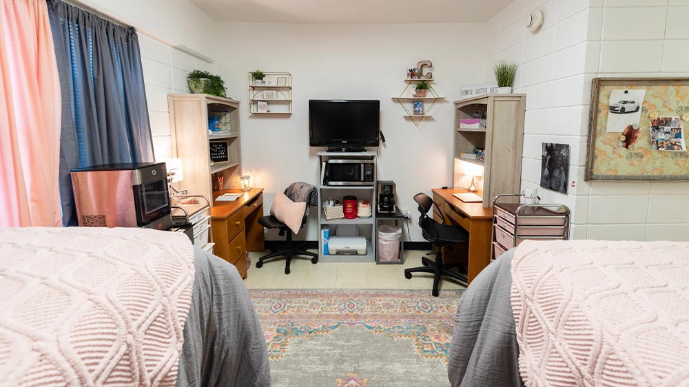 Bedroom layout showing two desks with chairs, shelving units, and a TV stand between them. Beds with pink and gray bedding are partially visible in the foreground.