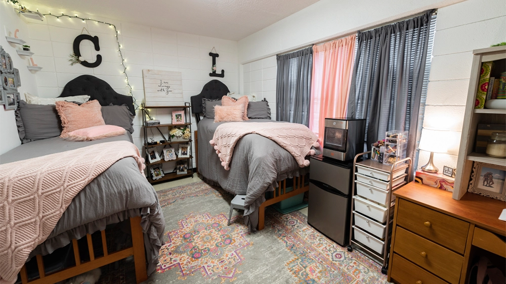 University Hall double bedroom decorated with gray bedding and pink accents. Two twin beds are arranged side by side with matching pillows and throws, a colorful area rug on the floor, and string lights framing the wall above photo collages and decorative letters.