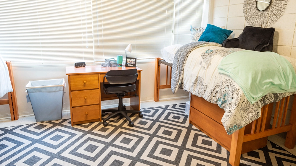 Close-up of a lofted bed with patterned bedding, a wooden desk and chair underneath, and a decorative wall mirror above the bed.