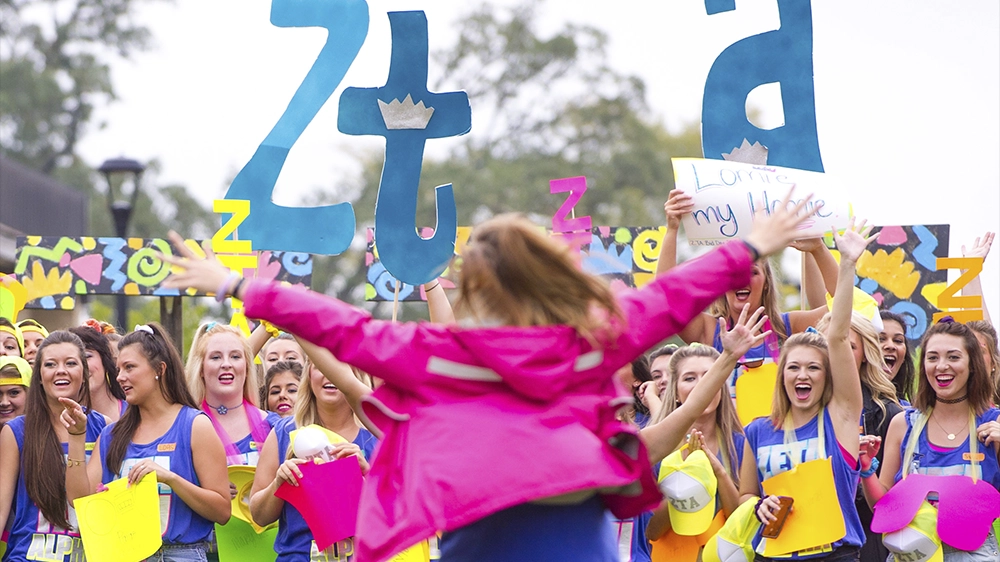 Group of people holding large blue Greek letters and colorful signs outdoors during Bid Day.