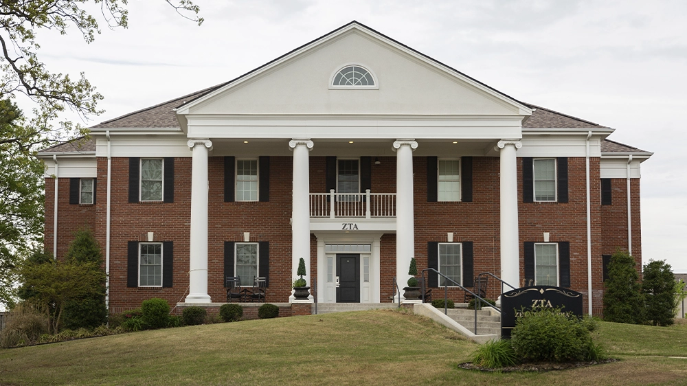 Two-story red brick sorority house with white columns, black shutters, and a triangular pediment. A black sign in front reads “ZTA.” The house sits on a grassy hill with shrubs along the walkway.