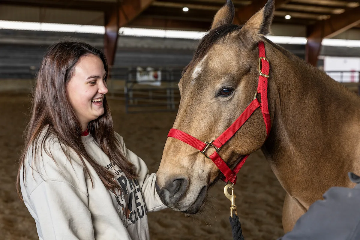 Smiling student pets a horse wearing a red halter inside an equine training arena.