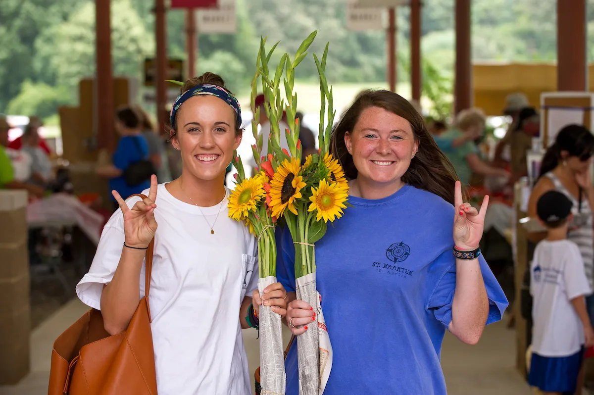 Two smiling students hold bouquets of sunflowers at an outdoor farmers market.
