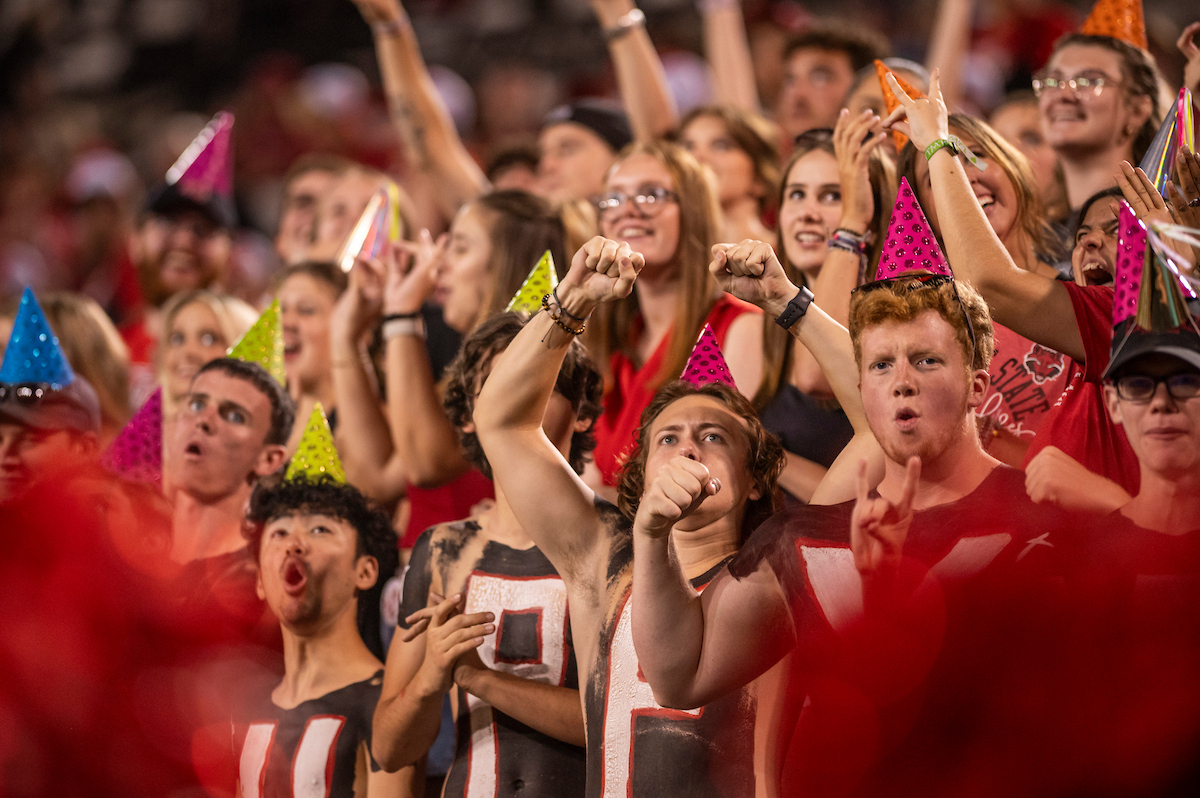 A-State student fans in party hats cheer during a game day celebration in the stands.