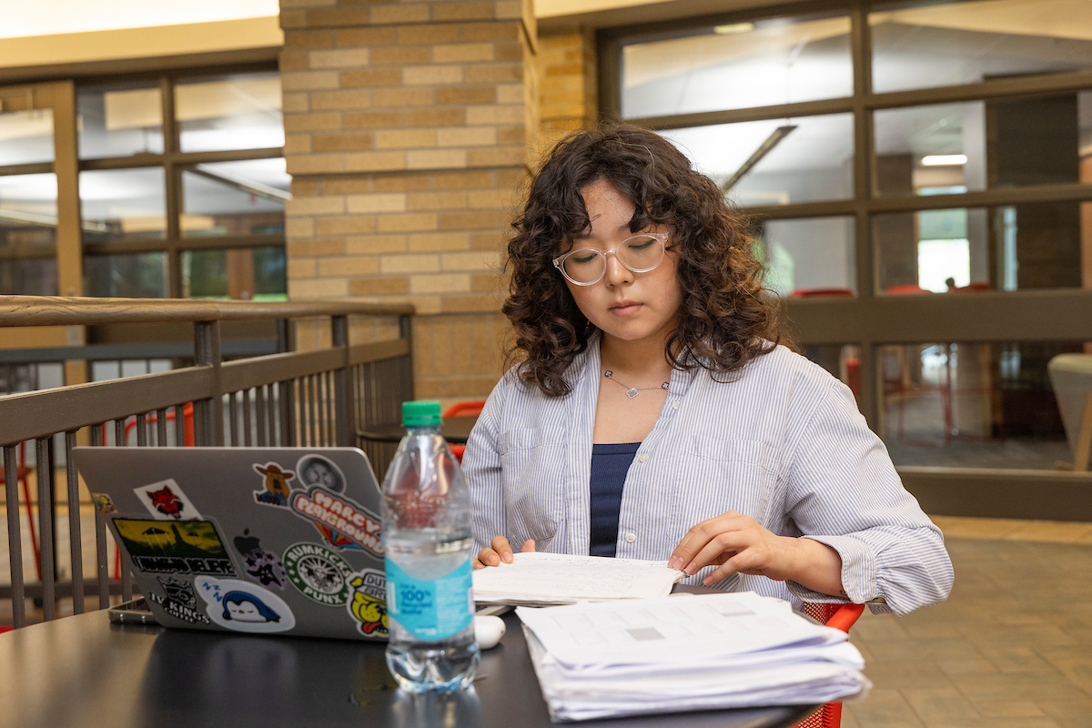An A-State student focuses on notes while studying with a laptop and papers at a table inside a campus building.