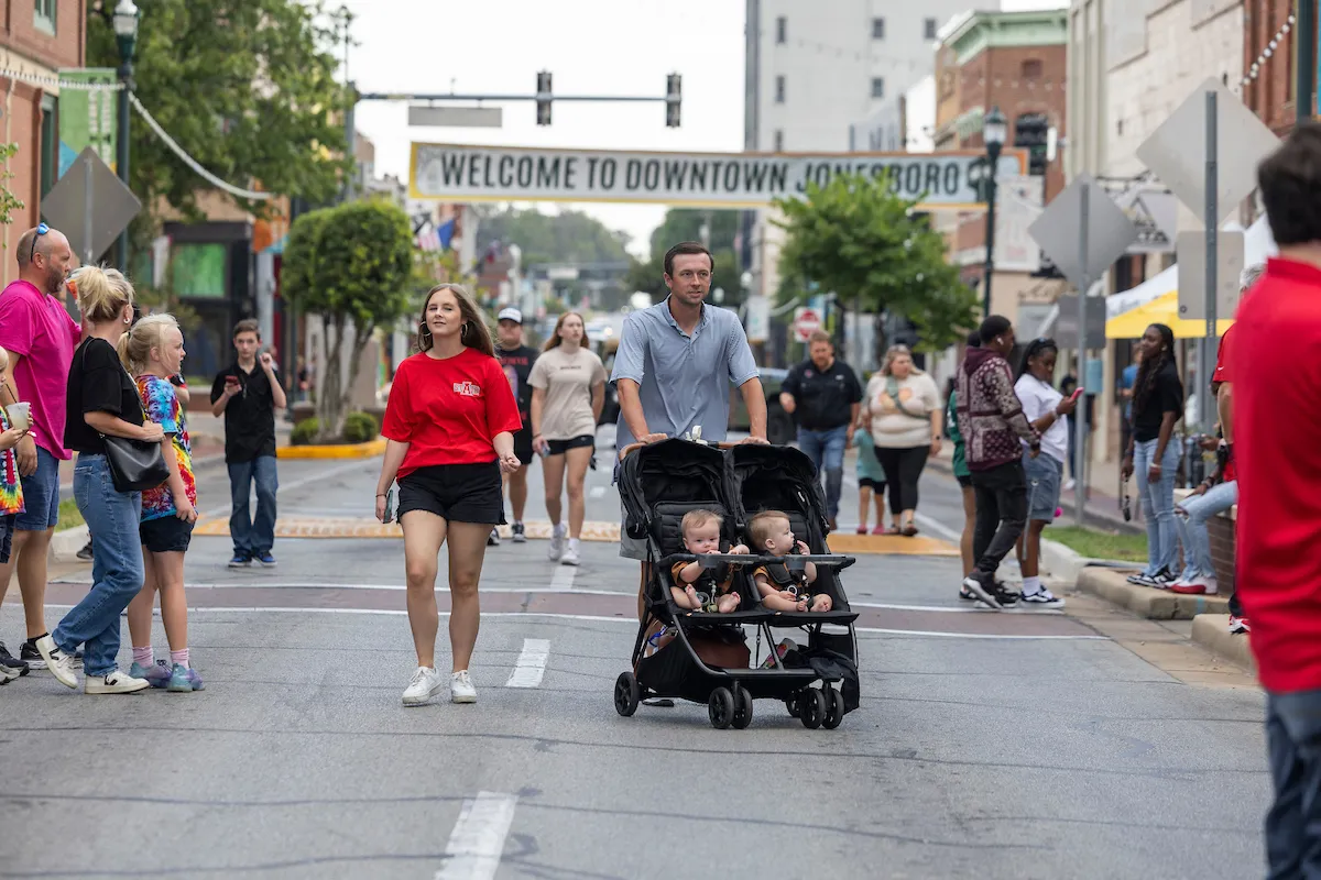 Families and students walk through a closed street during a community event in downtown Jonesboro under a welcome banner.